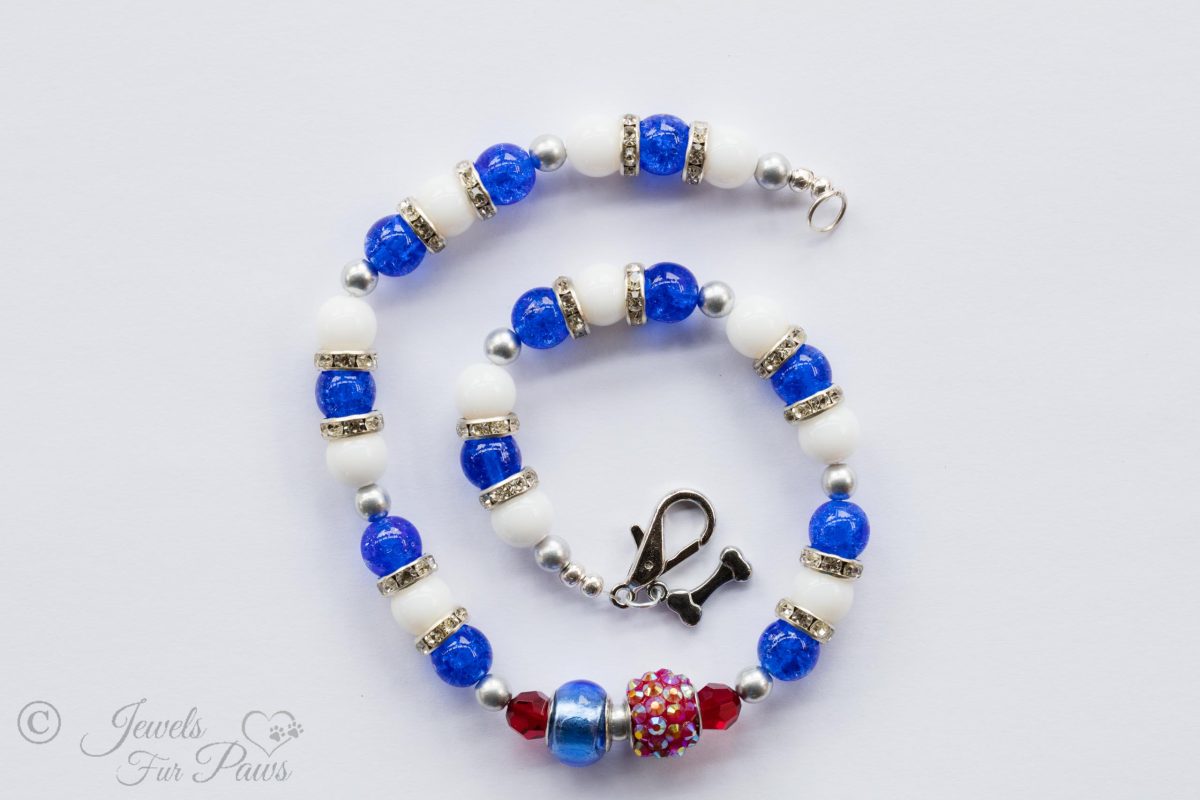white glass and blue glass beads with red crystals and a larger pave Egyptian rhinestone bead and channel set rhinestone rondell spacers and a hanging silver bone by the clasp on a white background