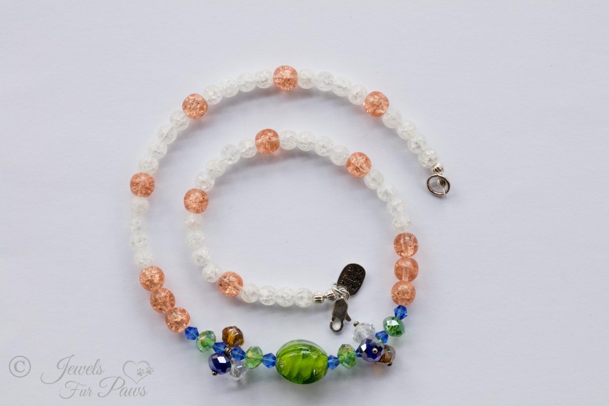 green lampwork glass beads surrounded by green and blue Swarovski crystals and crystal clusters complemented by peach and white cracked glass beads on white background