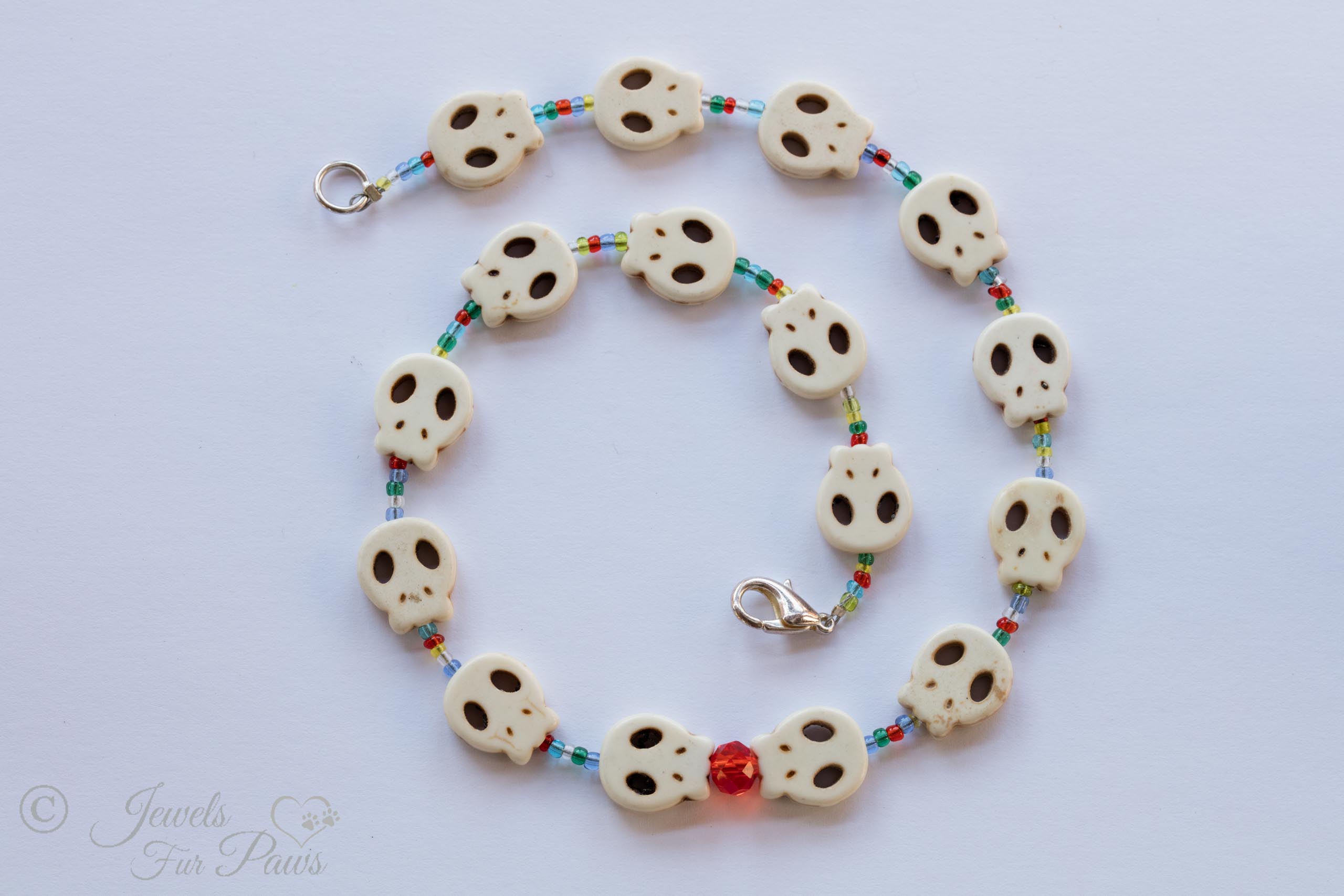 pet dog cat jewelry necklaces white skull charms strung with small blue, green, amber, orange spacer beads and feature a larger faceted red czech crystal on a white background