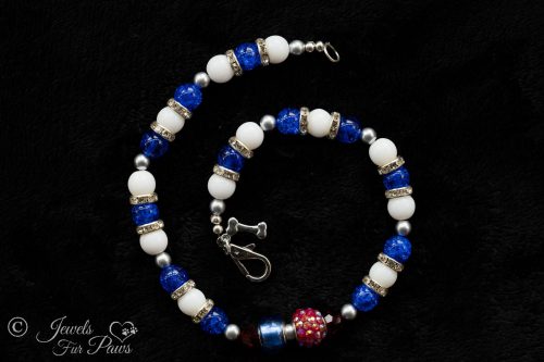 white glass and blue glass beads with red crystals and a larger pave Egyptian rhinestone bead and channel set rhinestone rondell spacers and a hanging silver bone by the clasp on a white background