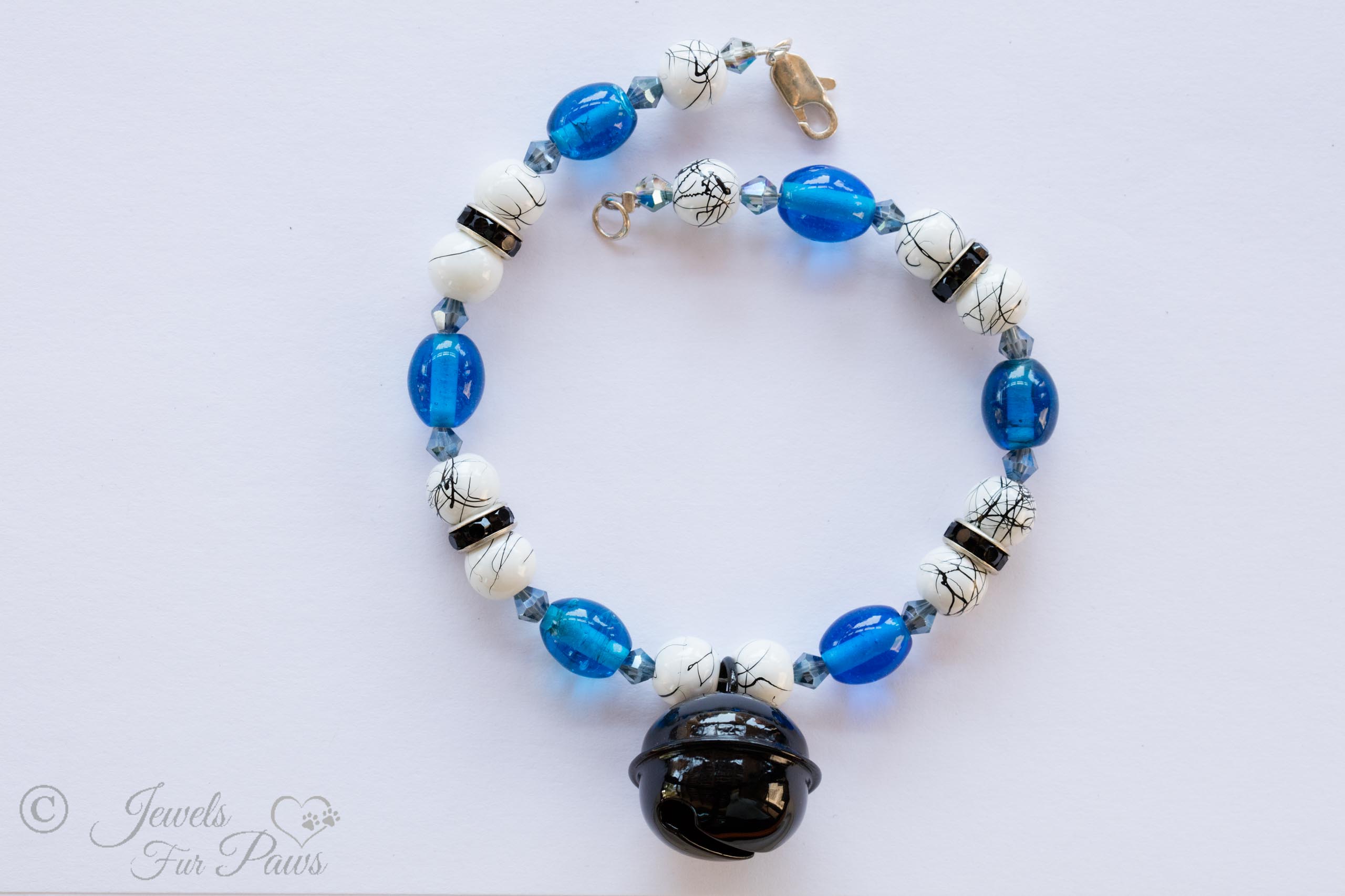 white and black marbleized beads with blue oval crystals and a hanging black bell on white background and black rhinestone spacers